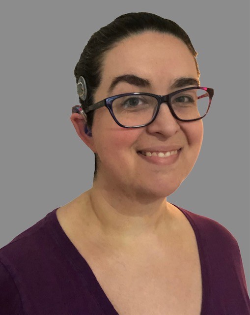 A smiling, young white woman with glasses wears her dark hair back to show her cochlear implant. 