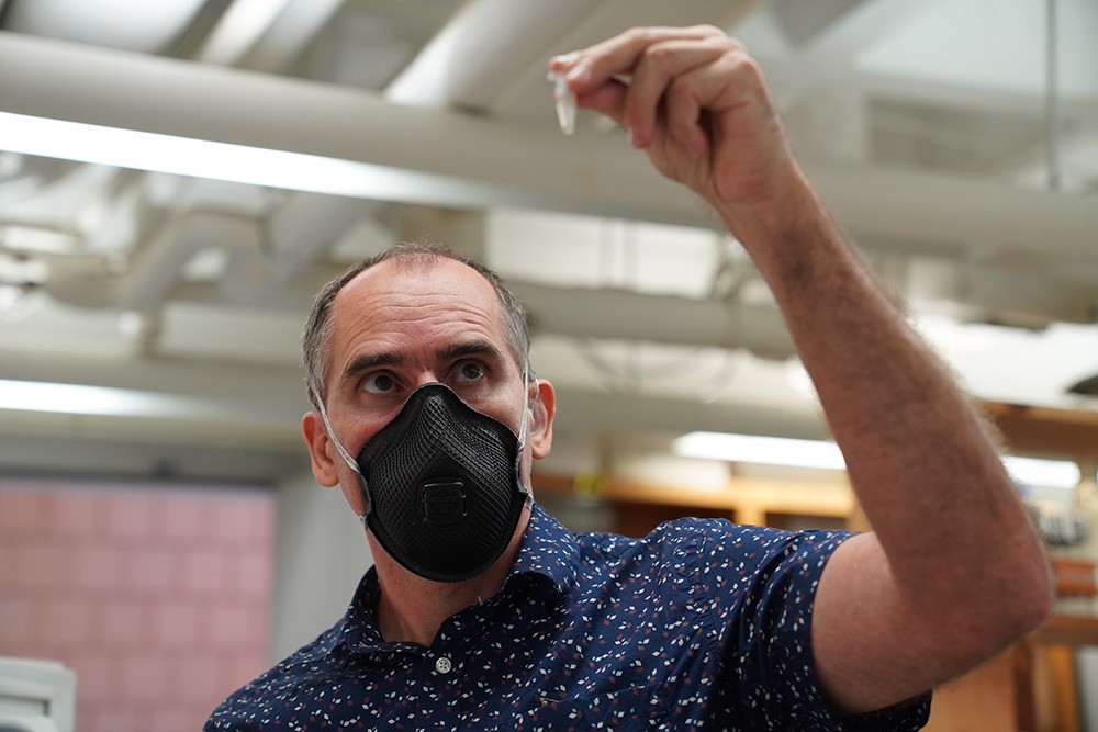 A white man with grey hair and a mask covering mouth and nose holds an eppendorf test tube and eyes its contents. He is wearing a dark blue shirt with a pattern and is in a laboratory type setting with white pipes in the background.